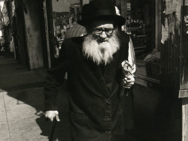 A black and white photograph captures an older man with a long white beard and a dark hat, walking with a cane. He holds a flower or similar object in his left hand and is casting a long shadow on the sunny sidewalk.